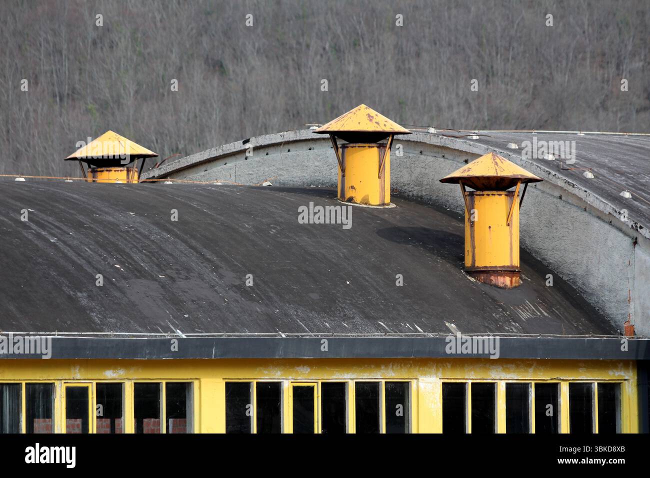 Tre sfiati in metallo giallo con tappi conici si trovano in cima a un tetto industriale nero curvo, con finestre con cornice gialla e uno sfondo di fronte senza fronzoli Foto Stock