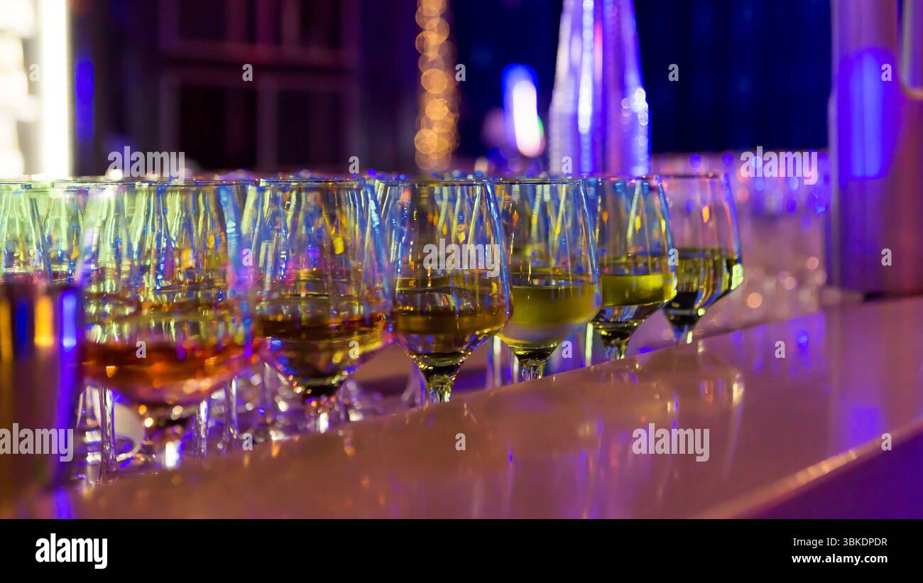 Una fila di bicchieri da vino pieni di bevande diverse, esposti su un bancone del bar con uno sfondo illuminato viola e blu. Foto Stock