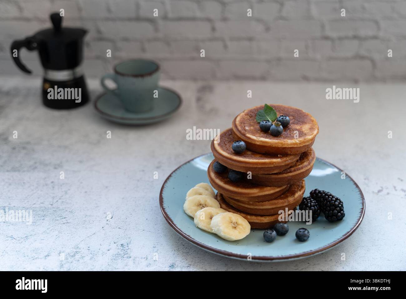 Gustosi pancake con frutti di bosco e fette di banana. Una moka e una tazza di caffè si trovano nelle vicinanze sulla superficie grigia, aggiungendo una calda atmosfera mattutina Foto Stock