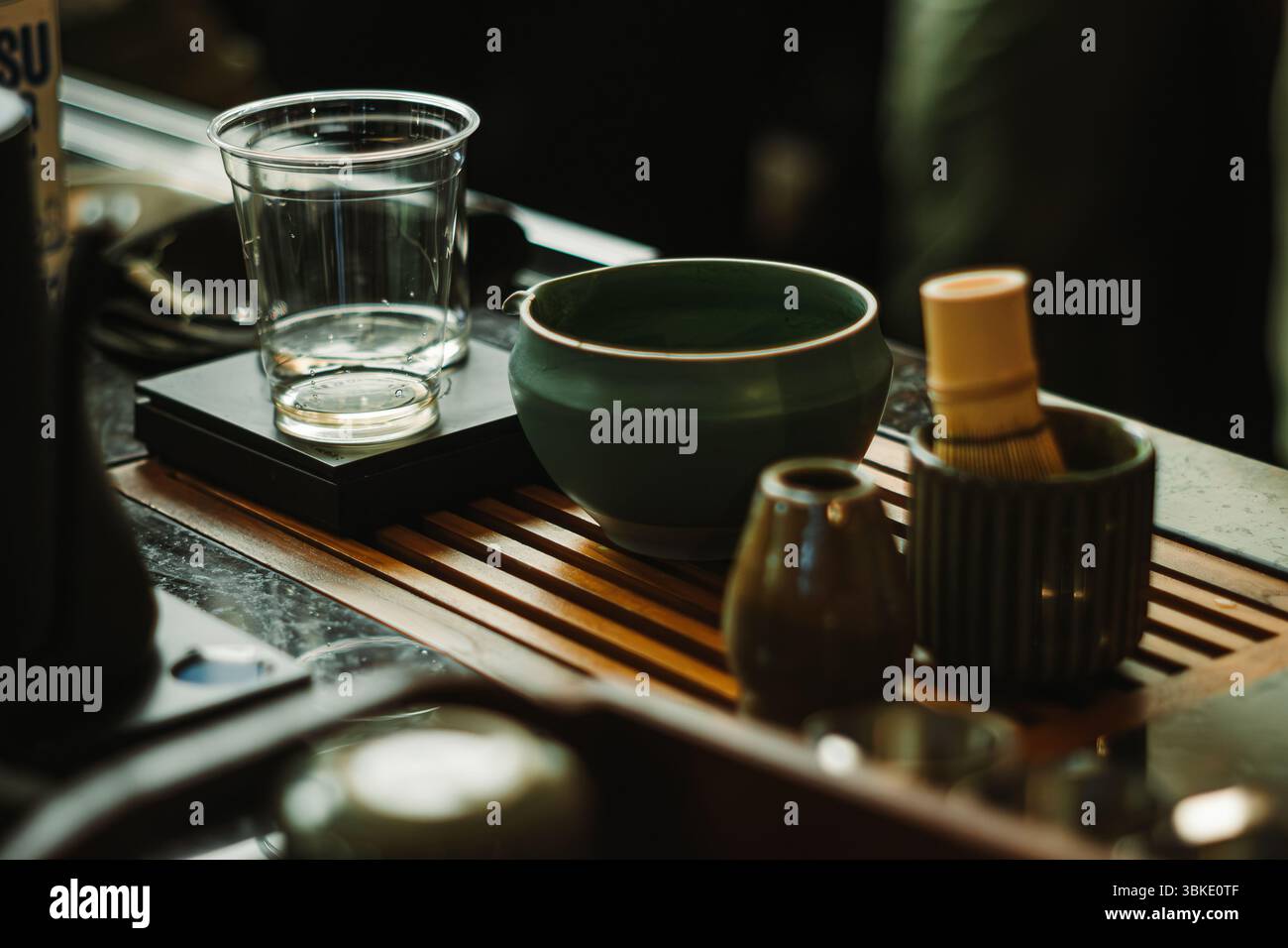 Primo piano di strumenti per il tè matcha su un vassoio di legno, tra cui un recipiente verde, una frusta di bambù, una tazza di ceramica, una caraffa e una tazza di plastica su una bilancia Foto Stock