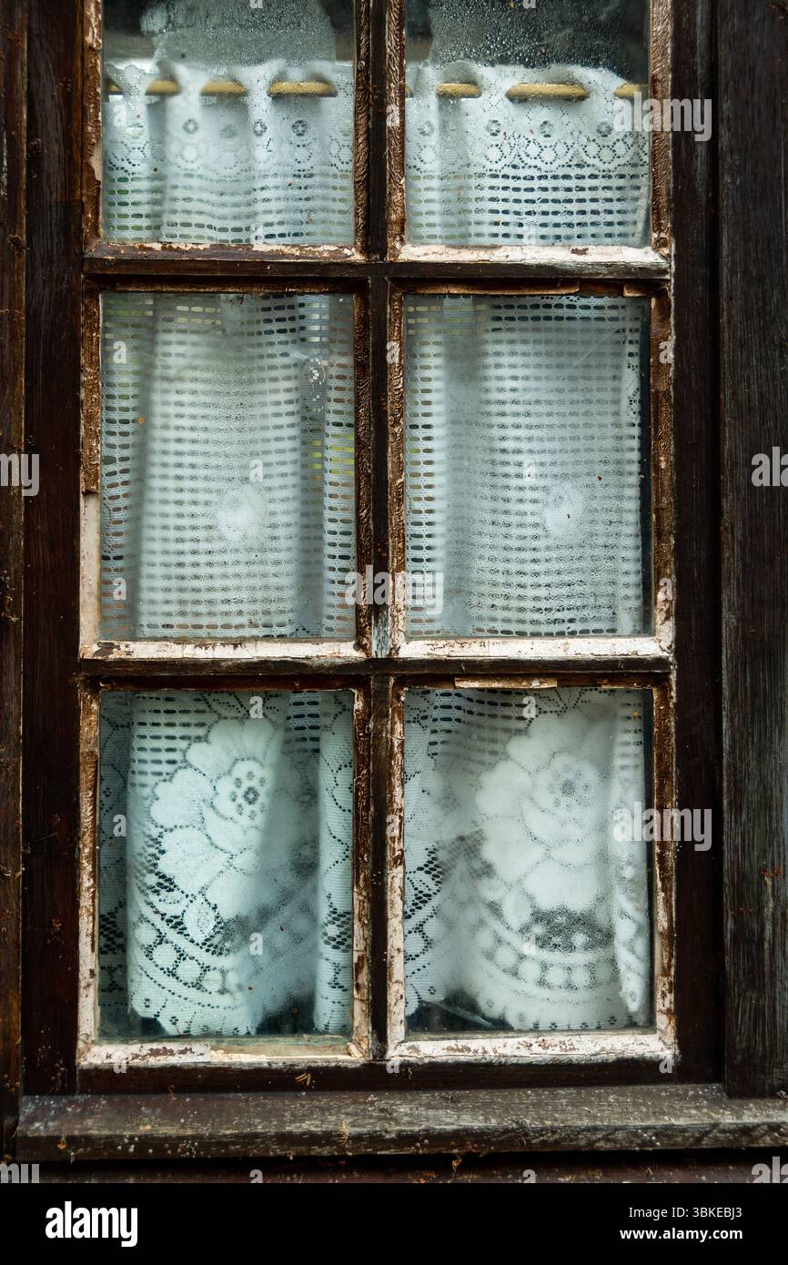 La finestra rustica mostra delicate tende in pizzo con intricati motivi floreali, leggermente smorzate dall'umidità esterna, Puy de Dome, Auvergne, Francia Foto Stock