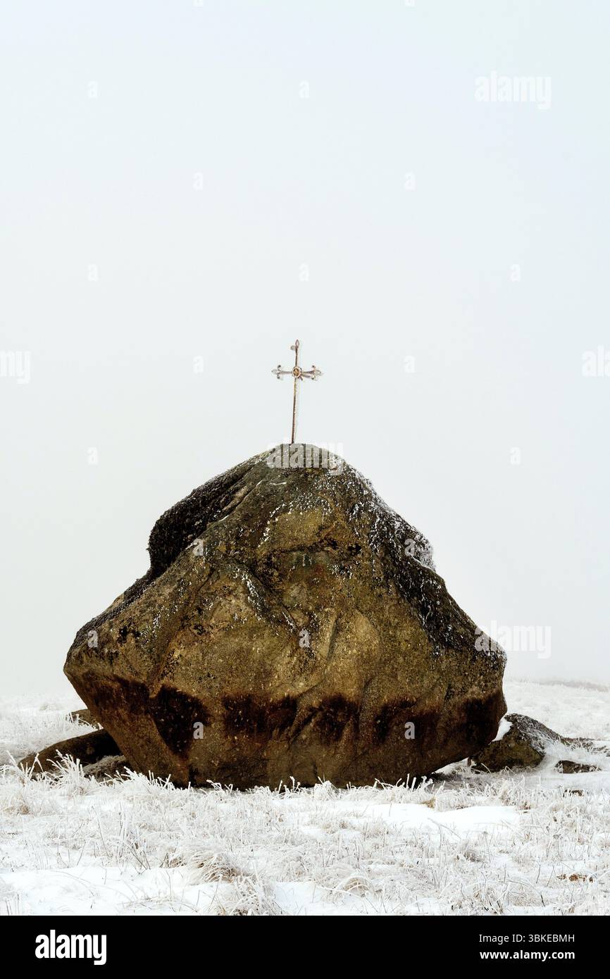 Una grande roccia sormontata da una croce si erge aspramente contro il paesaggio invernale dell'Altopiano Aubrac a Lozere, Occitanie, in Francia Foto Stock