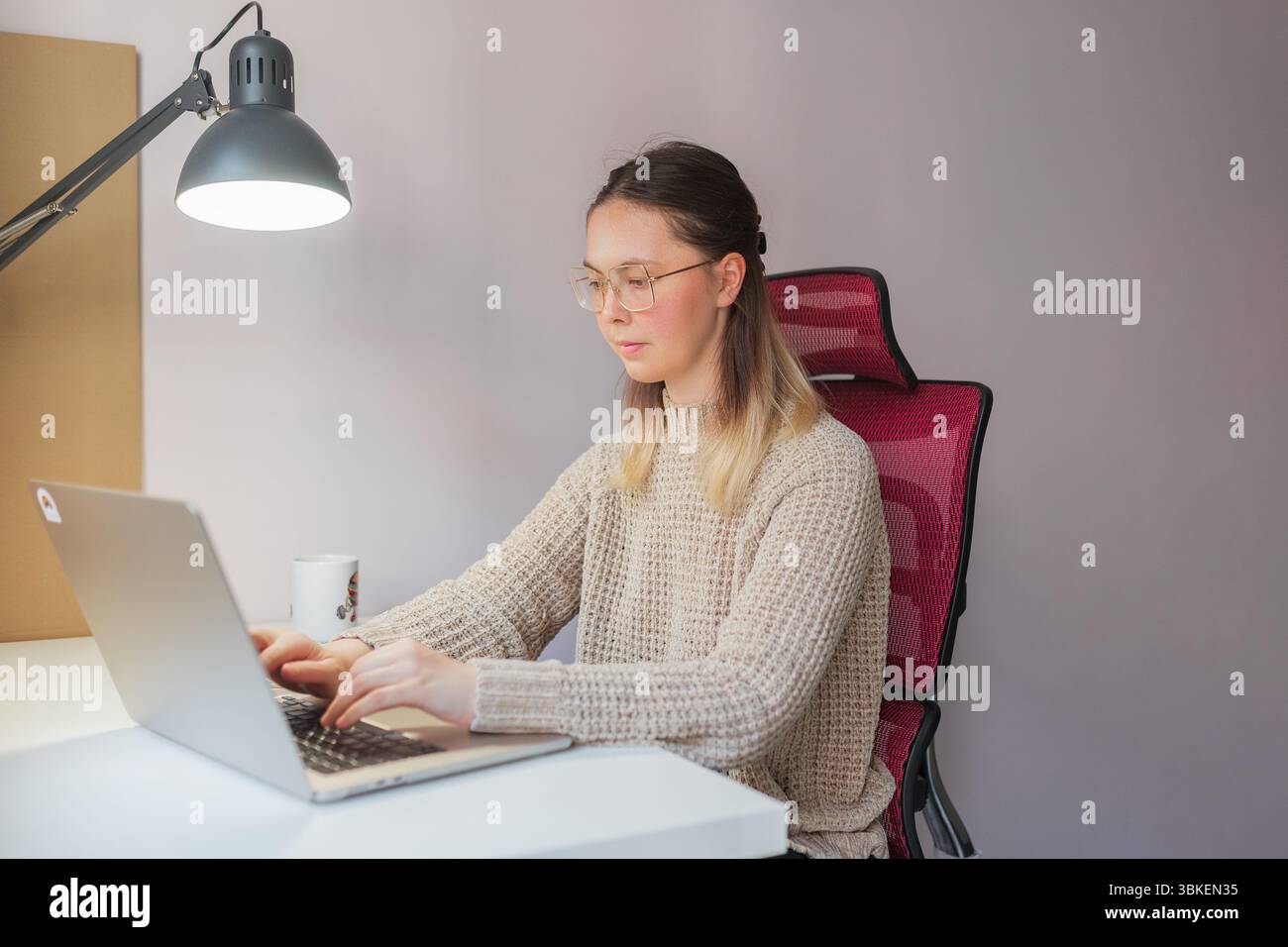 Donna che scrive su un computer portatile sotto la lampada da scrivania nell'ufficio domestico con sedia in rete rossa. Lavoratrice libera seduta alla scrivania bianca, indossando un maglione beige Foto Stock