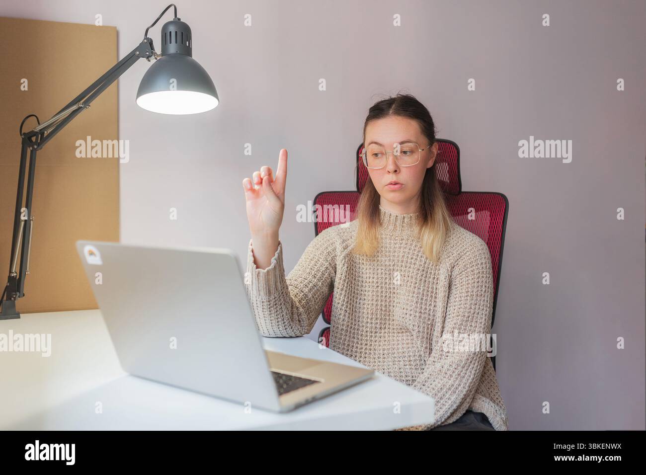 Donna che alza il dito, sembra spiegare qualcosa durante la videochiamata. Gesto sicuro e look serio, giovane donna che interagisce tramite notebook, in interni Foto Stock