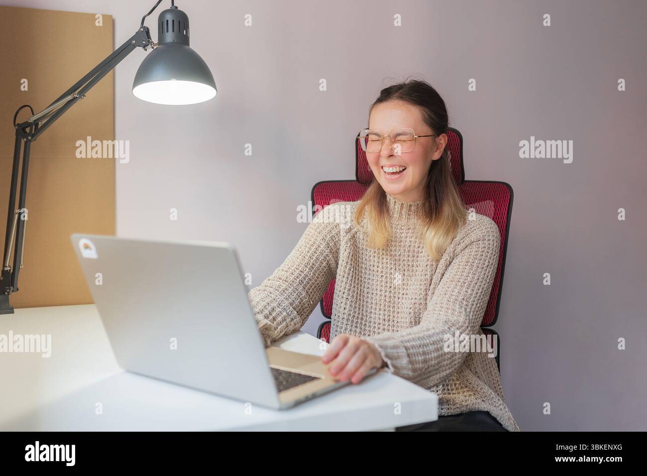 Donna sorridente con gli occhi chiusi per piacere mentre è seduta al notebook. L'emozione gioiosa catturata come donna in un ufficio domestico informale reagisce a qualcosa sullo schermo Foto Stock