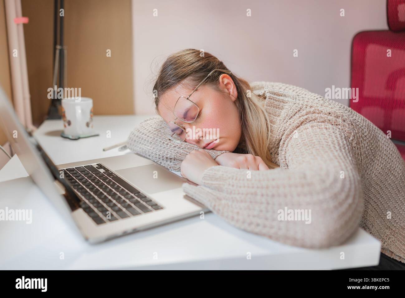 Donna stanca che dorme alla scrivania, mano sul laptop, poggiatesta. Comoda sistemazione in casa con donna che dorme sulla scrivania bianca, occhiali inclinati e. Foto Stock