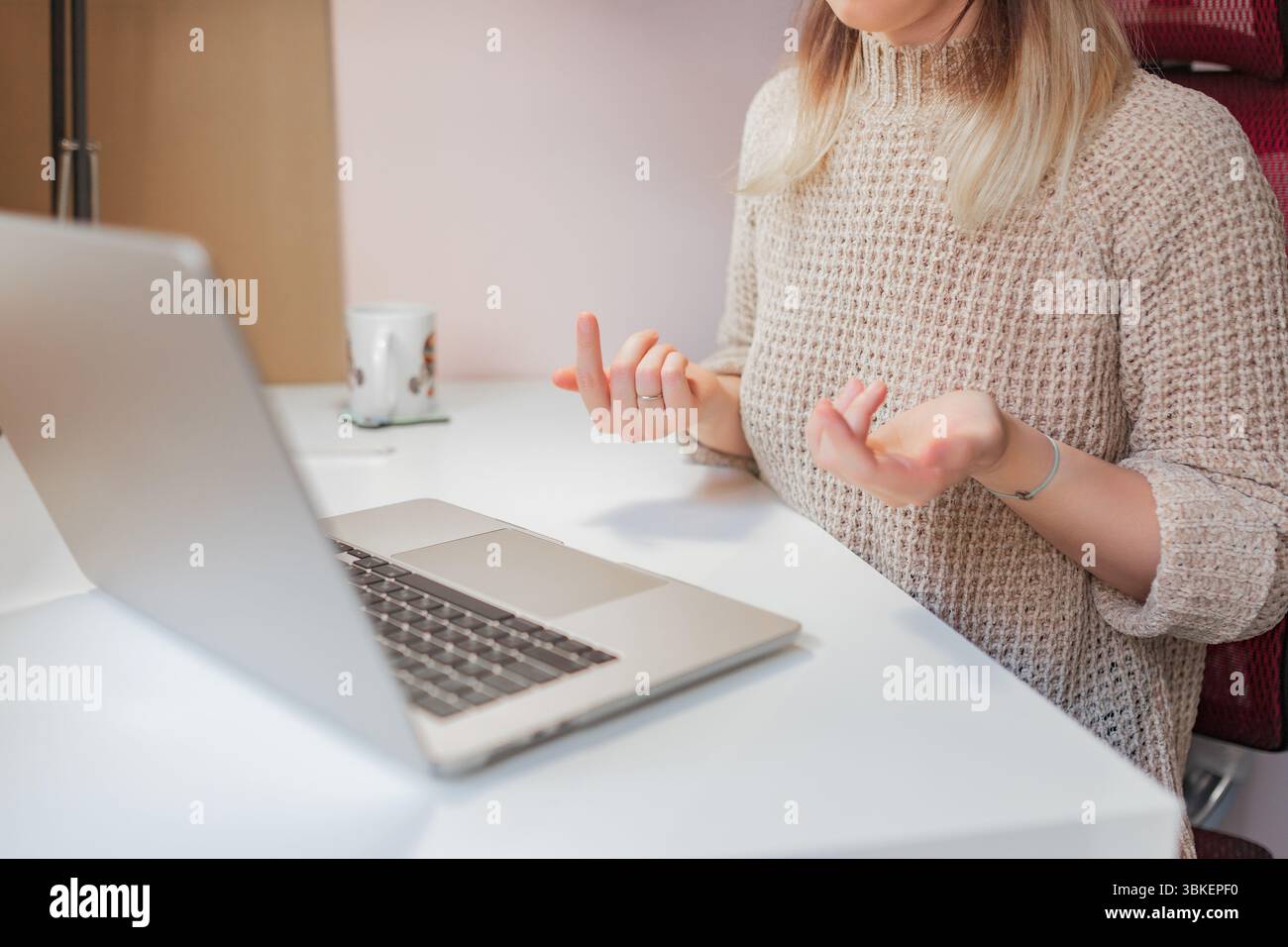 Una donna si toglie durante una riunione remota mentre è seduta al computer portatile in casa. Mani leggermente sollevate con palmi interni, la donna esprime incertezza o truffa Foto Stock