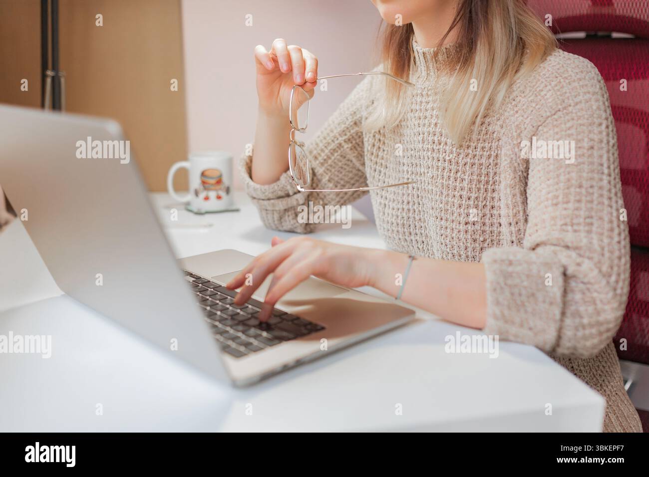 La donna solleva gli occhiali mentre lavora con un computer portatile durante le sessioni di lavoro in casa. La signora regola gli occhiali mentre digita il laptop a casa, indossa un abbigliamento accogliente e calmo Foto Stock