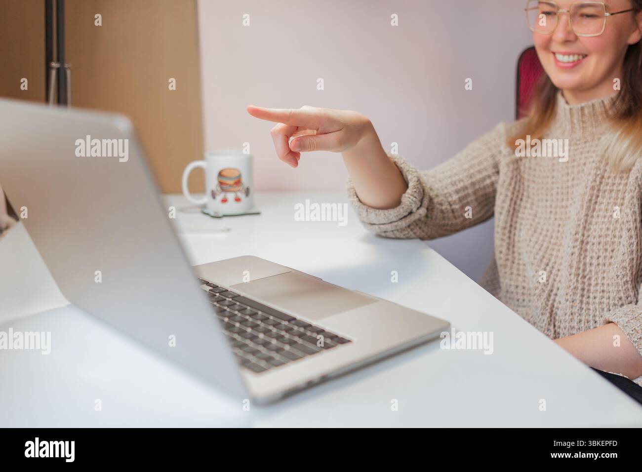 La donna sorride e punta sullo schermo durante una divertente videochiamata. Scena luminosa in interni con donna sorridente che punta il dito verso lo schermo di un notebook, casual Foto Stock