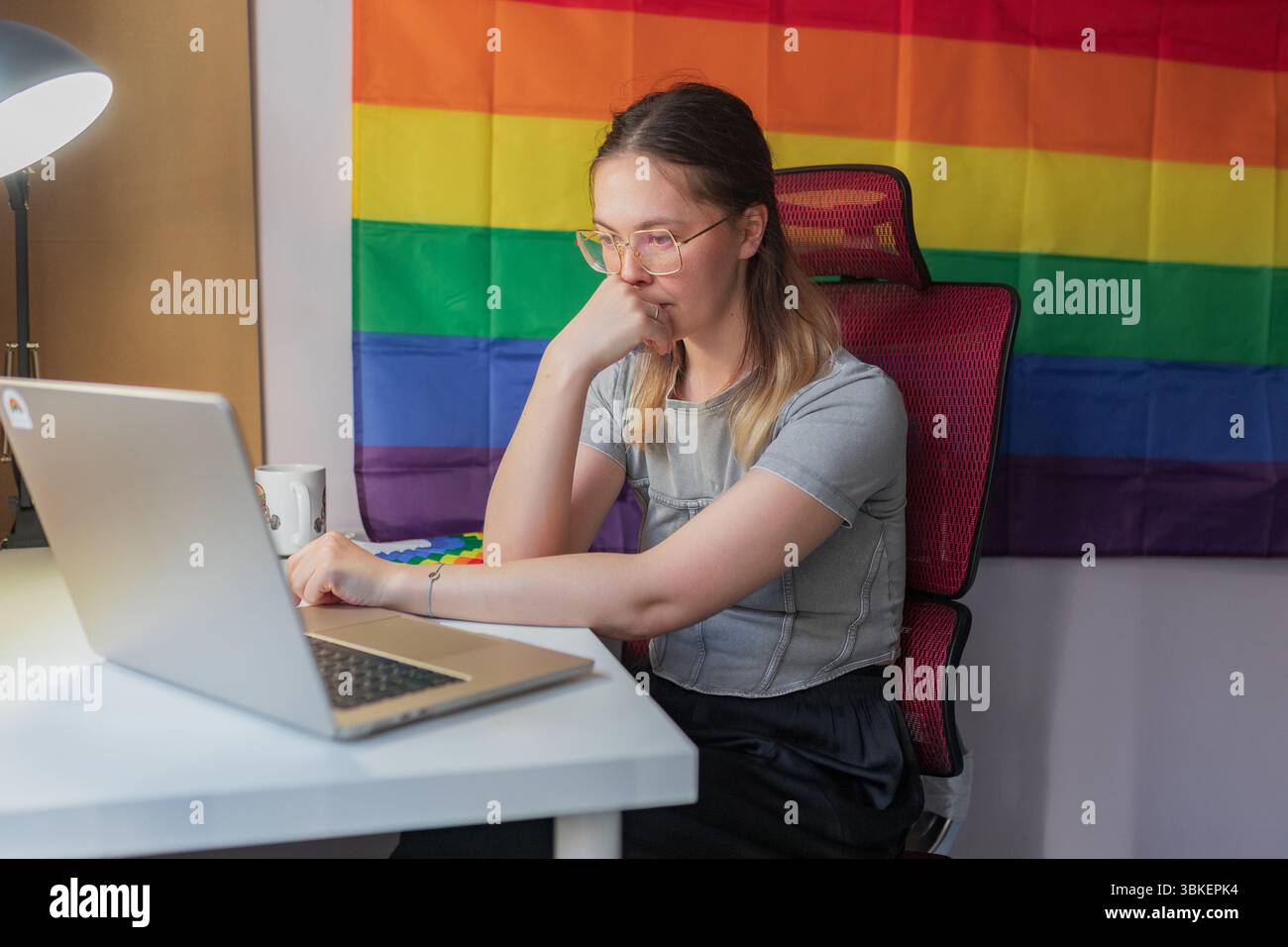 Una donna concentrata si appoggia la mano sul viso mentre lavora da remoto in una stanza a tema orgoglio. Una giovane donna professionista si siede con cura davanti a un notebook con mento Foto Stock