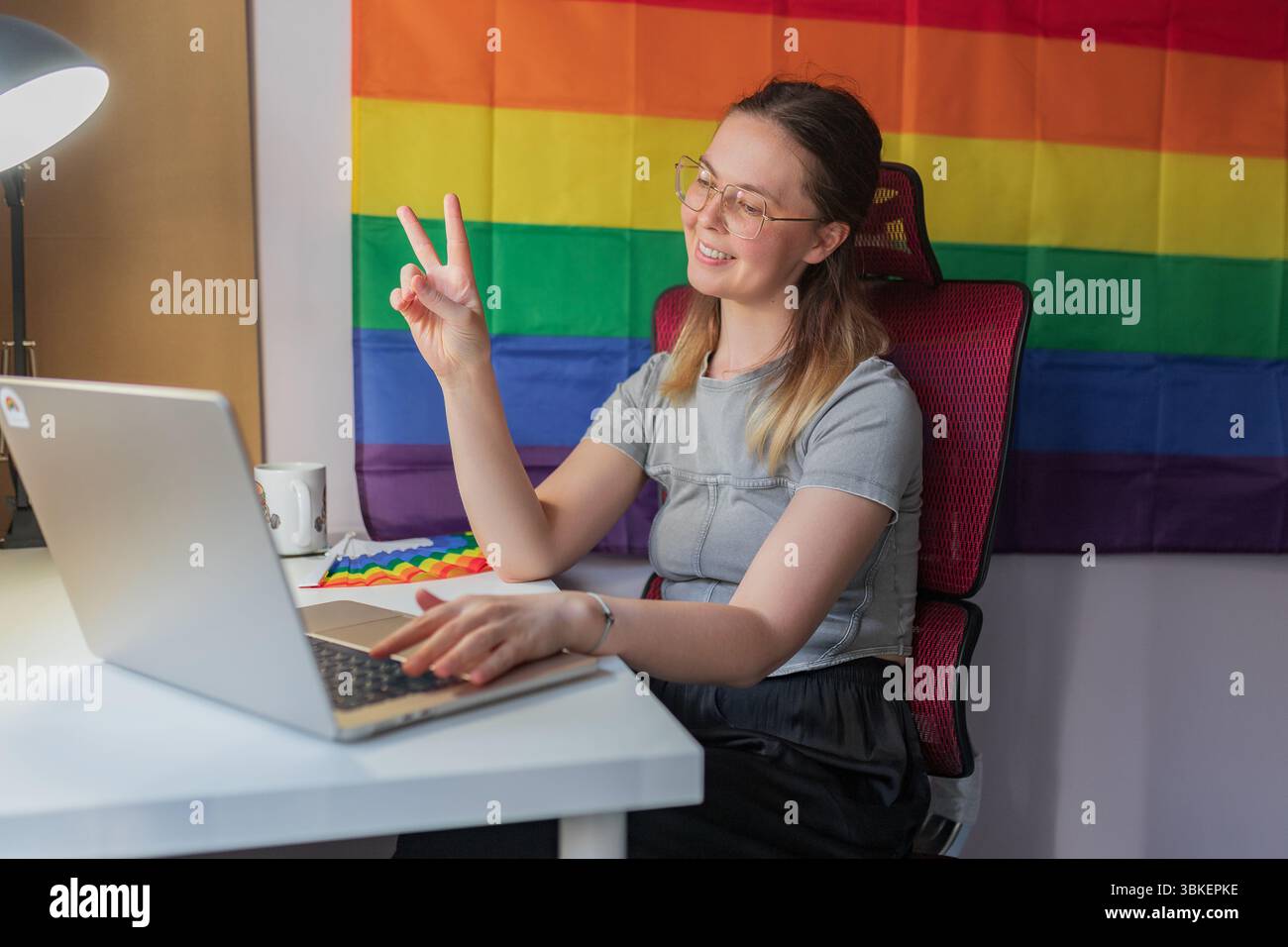 Donna sorridente che mostra un segno di pace durante la videochiamata, sfondo arcobaleno. Donna con gli occhiali seduti sullo spazio di lavoro di casa che mostra un segno di pace, colorata Foto Stock