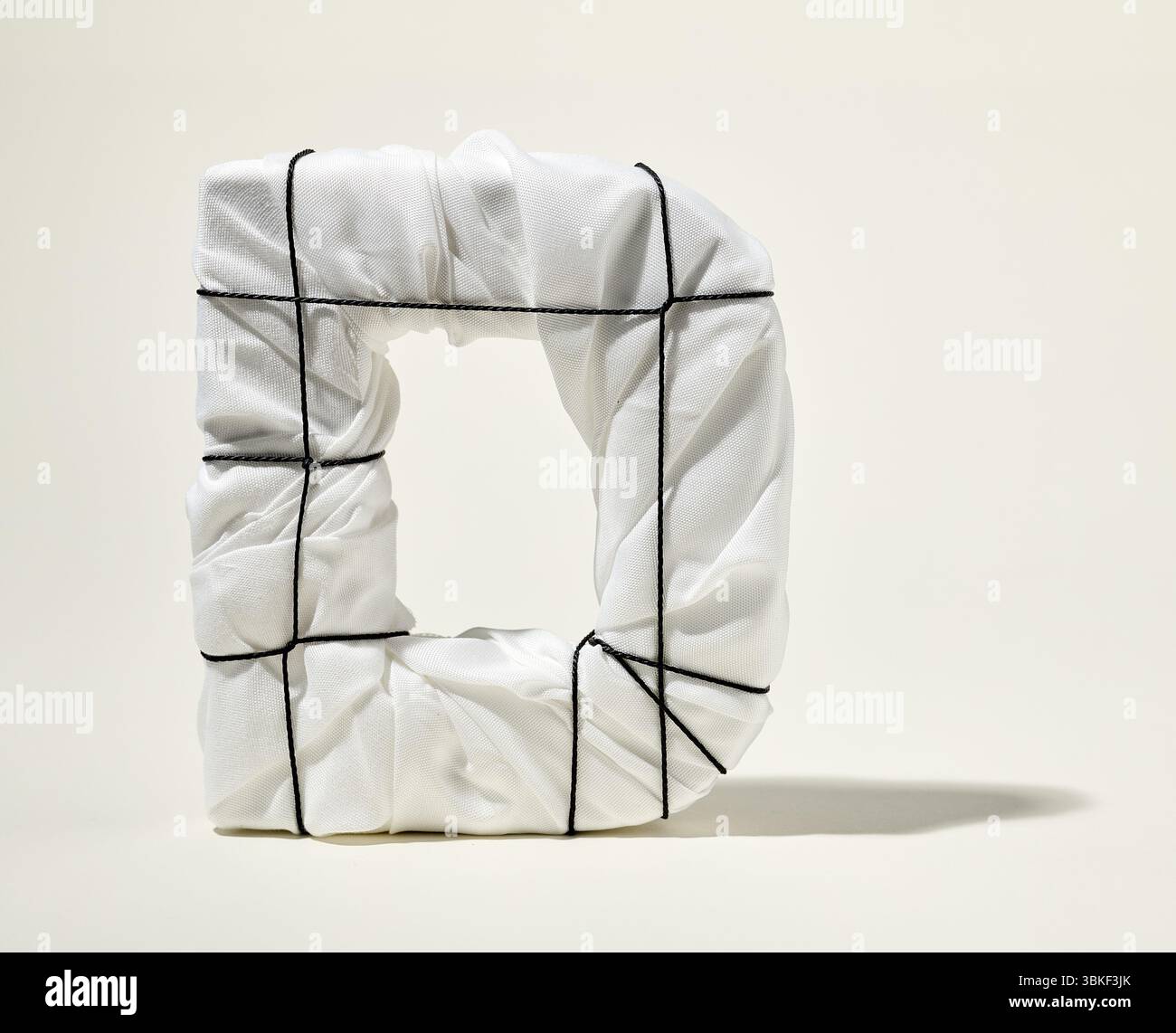 Lettera D avvolta in tessuto bianco con cordoncino nero su sfondo beige Foto Stock