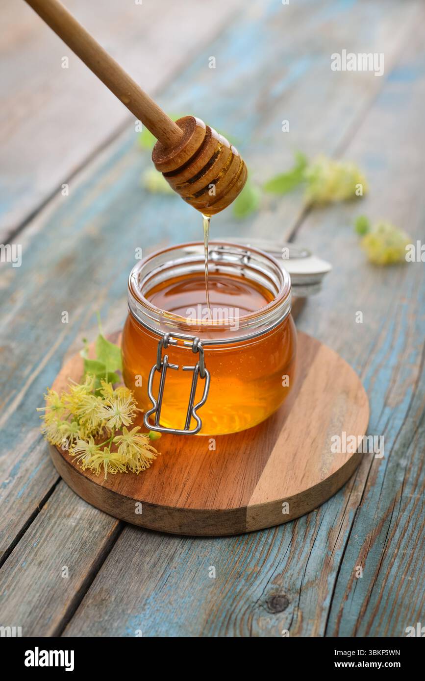 Miele di tiglio in vaso di vetro con dipper di miele di legno e fiori di tiglio su fondo di legno Foto Stock