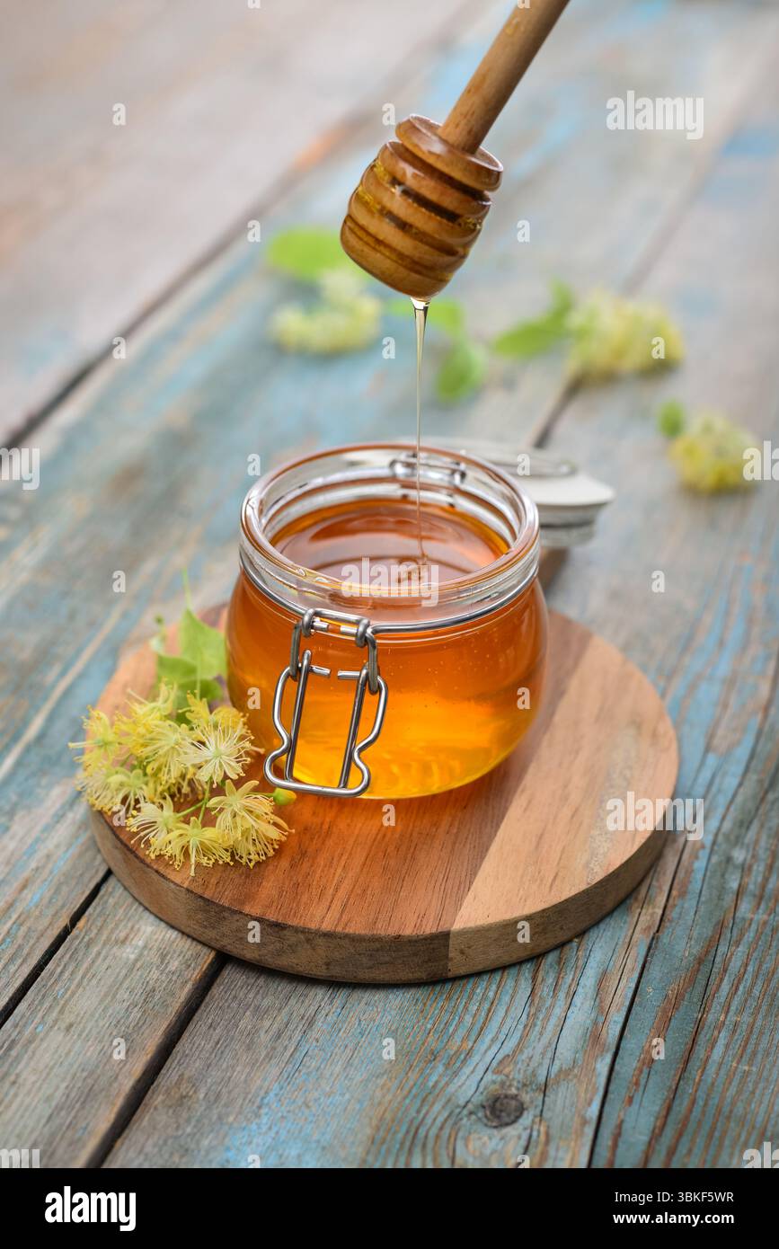 Miele di tiglio in vaso di vetro con dipper di miele di legno e fiori di tiglio su fondo di legno Foto Stock