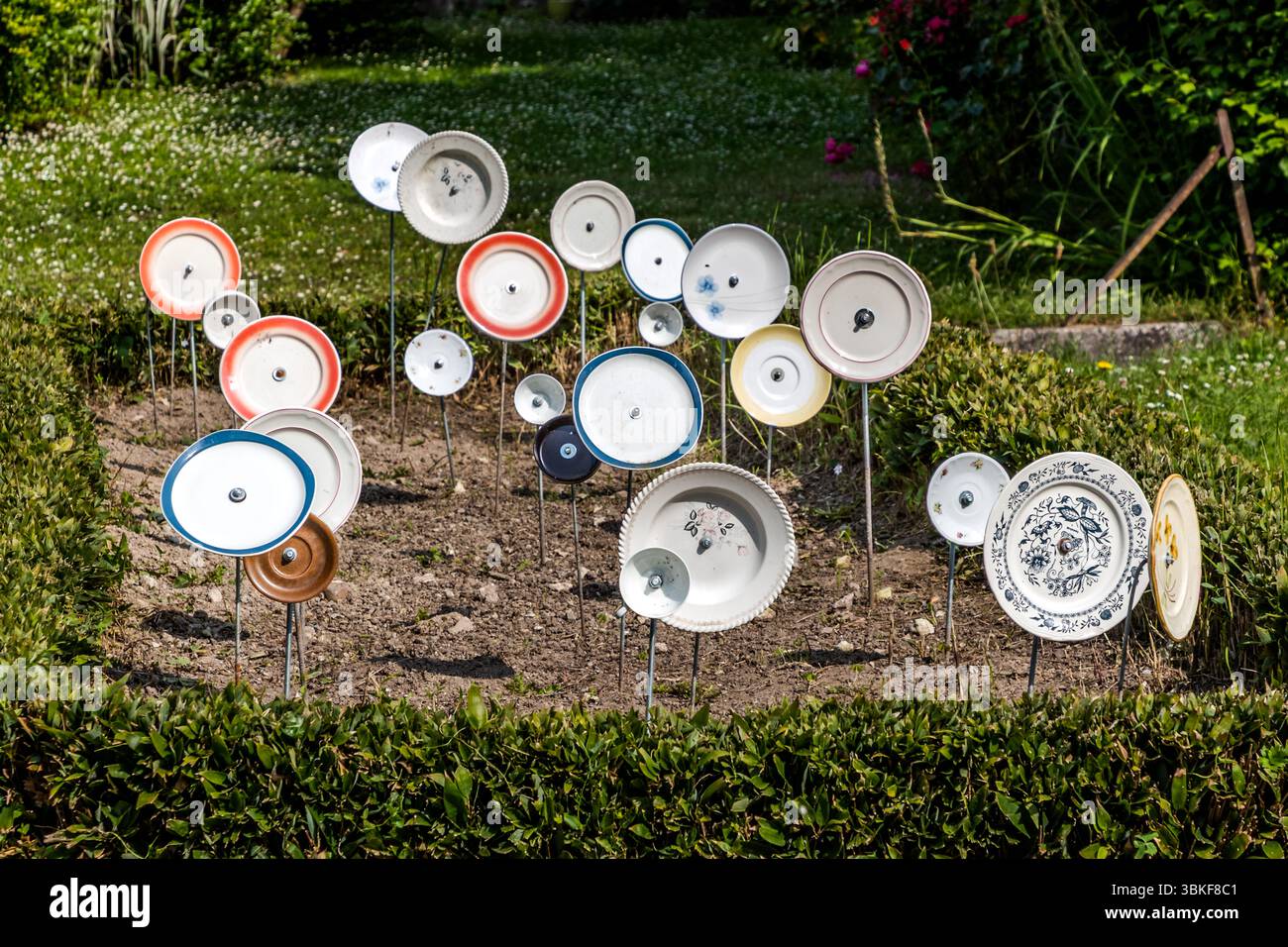 Piatti su pali in un giardino frontale in Francia, Nouvelle-Aquitaine. Il motivo si inserisce nel proverbio tedesco "Über den Tellerrand schauen" (guarda oltre il bordo del piatto), che si riferisce a un atteggiamento aperto e curioso in cui si è disposti a trascendere i propri confini e ad imparare cose nuove. La frase è spesso usata in connessione con il cosmopolitismo, un cambiamento di prospettiva o innovazione. Pensare fuori dagli schemi. L'installazione decorativa del giardino comprende vari pezzi di stoviglie attaccati ai bastoncini e collocati in un letto di fiori per creare una scena giocosa e artistica. L'installazione è Foto Stock