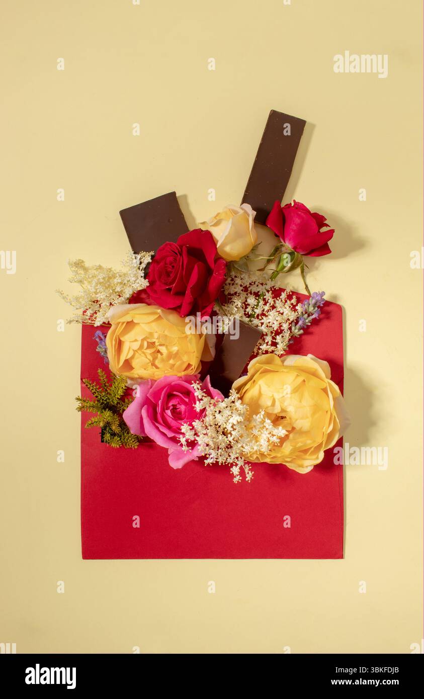Composizione creativa composta da fiori colorati e cioccolato fondente con busta rossa su sfondo beige. Disposizione piatta, vista dall'alto, spazio di copia Foto Stock