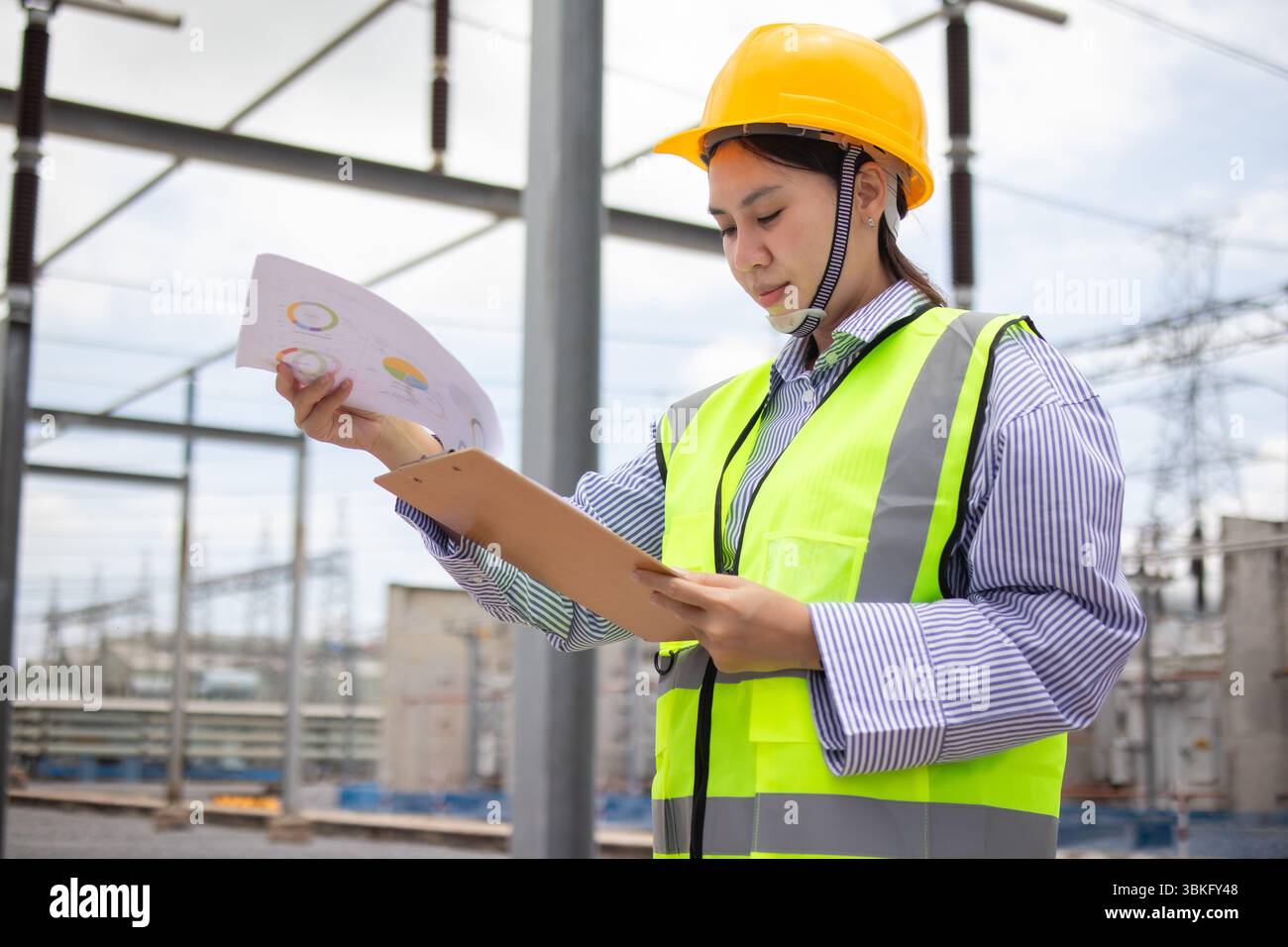 Un ingegnere dell'energia sta lavorando in una sottostazione, esaminando i documenti con particolare attenzione. il membro del personale indossa elmetto giallo e giubbotto riflettente, ensurin Foto Stock