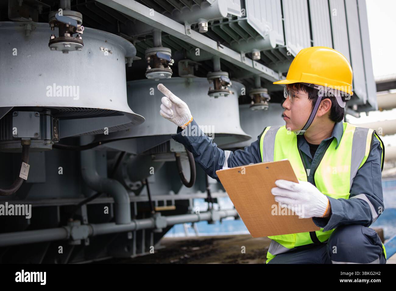 Un tecnico che indossa un elmetto giallo esegue l'ispezione del sistema di raffreddamento su un grande trasformatore presso una sottostazione esterna, garantendo sicurezza e funzionamento dell'eff Foto Stock