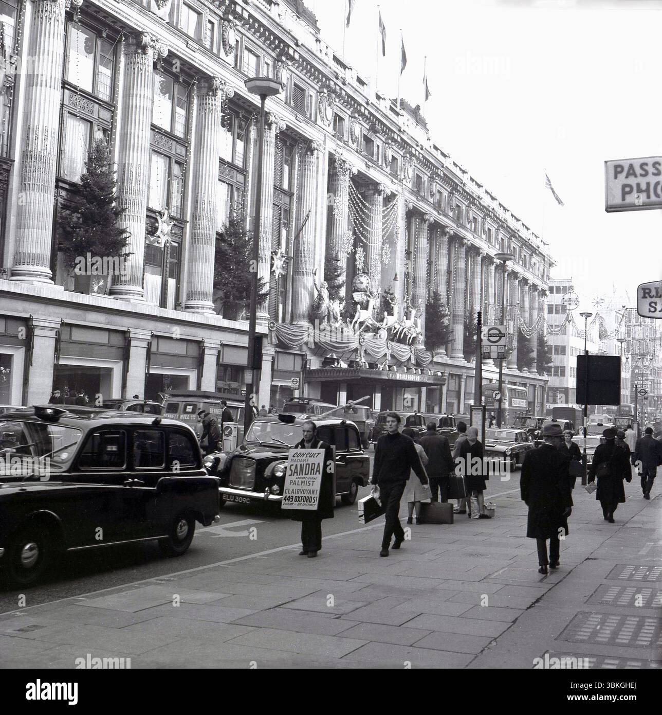 1966, periodo storico, natalizio e una trafficata Oxford Street fuori dal grande magazzino Selfridges con negozi e taxi. Sul marciapiede un uomo con una tavola di sabbia che pubblicizza "Madame Sandra", il principale palmista e chiaroveggente di Londra al 449 di Oxford St, di fronte a Selfridges. Foto Stock