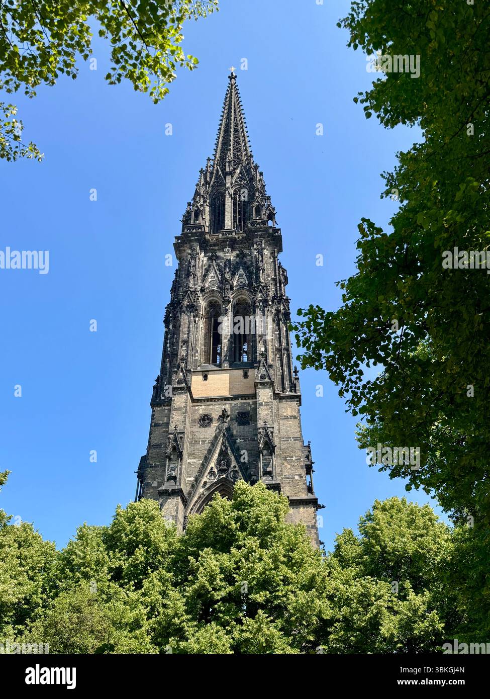 Foto della chiesa commemorativa di San Nicola ad Amburgo, Germania, con la sua alta guglia gotica incorniciata da alberi verdi in una luminosa giornata estiva. - Immagine stock catturata con smartphone