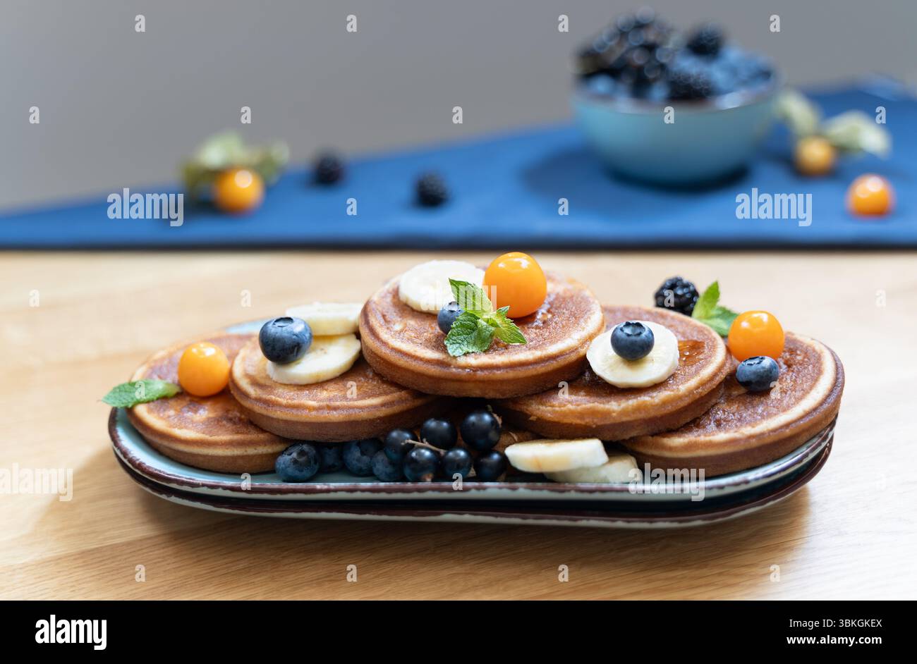Pancake con piatti blu su un tavolo di legno e frutti di bosco sullo sfondo, la calda luce del giorno dall'alto aggiunge un tocco di luce Foto Stock