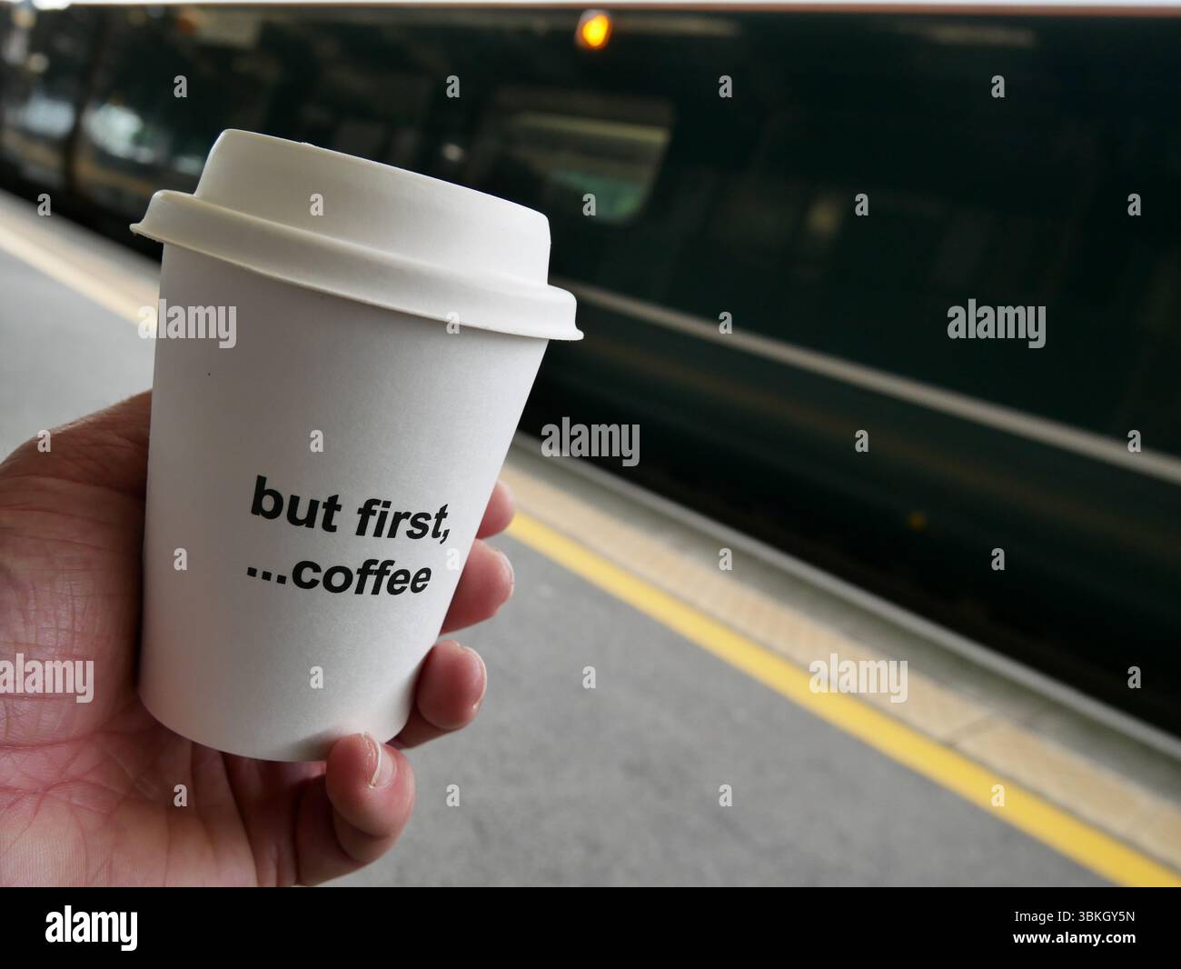 POV mano che tiene una tazza da asporto sul binario della stazione ferroviaria bevendo un caffè mattutino durante gli spostamenti in attesa di un treno. REGNO UNITO Foto Stock