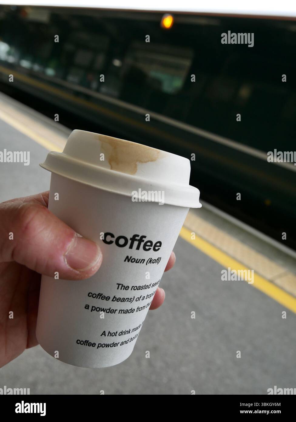 POV mano che tiene una tazza da asporto sul binario della stazione ferroviaria bevendo un caffè mattutino durante gli spostamenti in attesa di un treno. REGNO UNITO Foto Stock