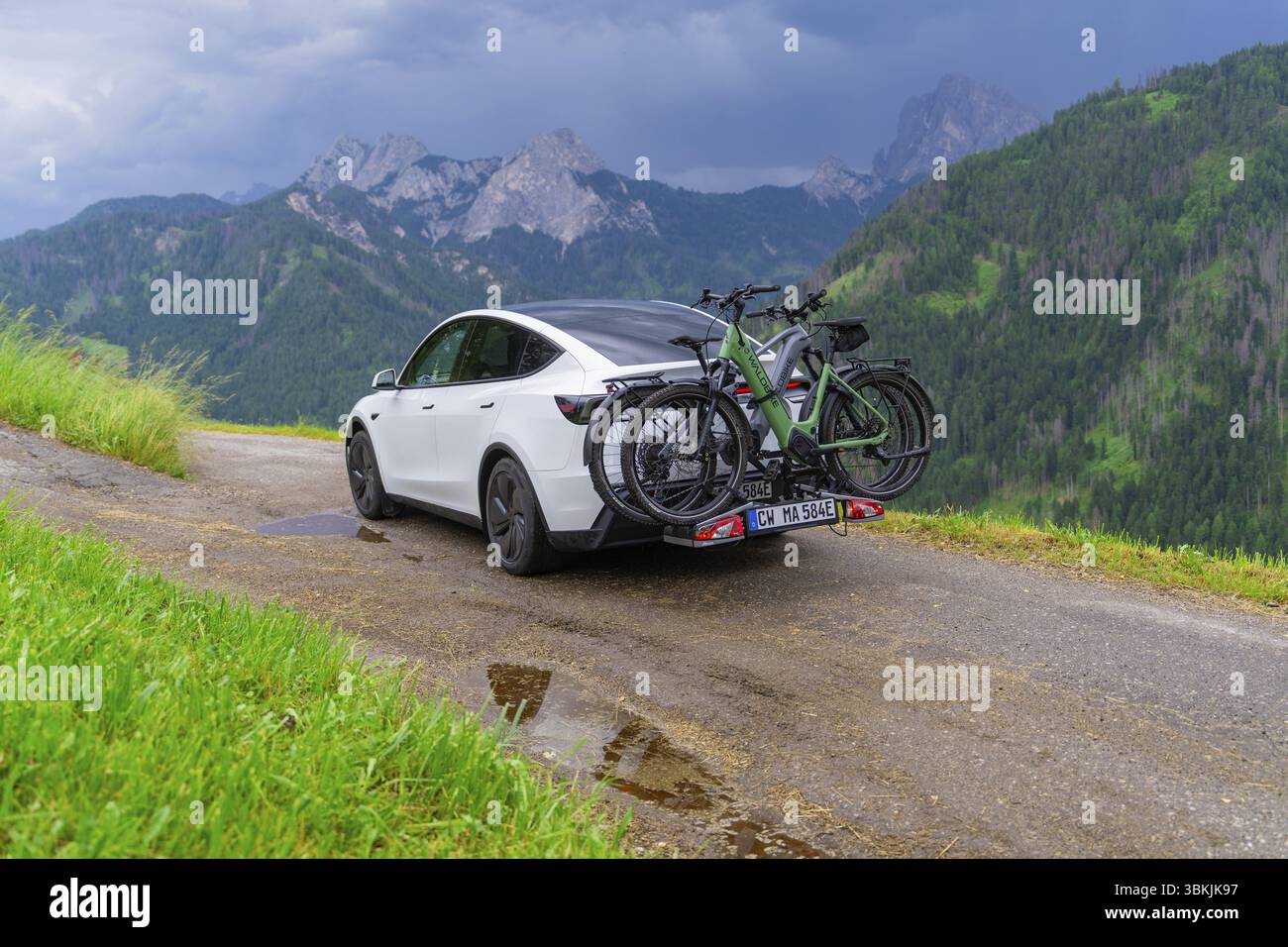 Auto bianca con biciclette su strada tortuosa, immersa nel verde del paesaggio montano sotto il cielo buio, auto elettrica in vacanza, Dolomiti, alto Adige, Dolom Foto Stock