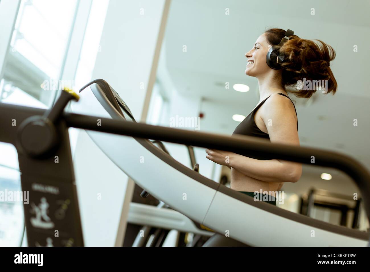 Un atleta allegro corre su un tapis roulant, immerso nella musica, in una palestra luminosa durante le ore diurne, abbracciando il fitness con gioia. Foto Stock