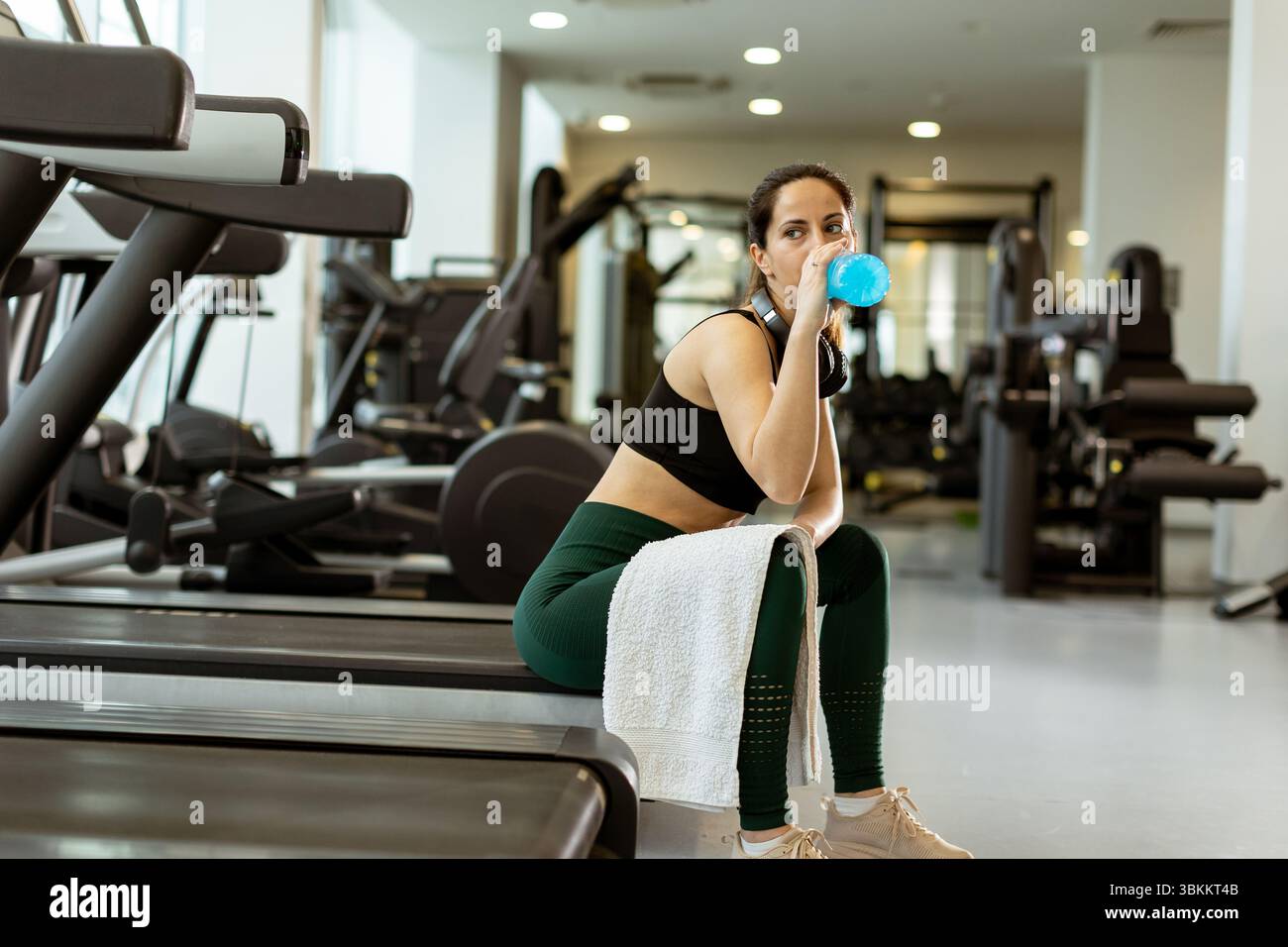 Una donna siede su una panca da palestra, sorseggiando una bevanda azzurra dopo un intenso allenamento, circondata da attrezzature fitness in una struttura moderna. Foto Stock