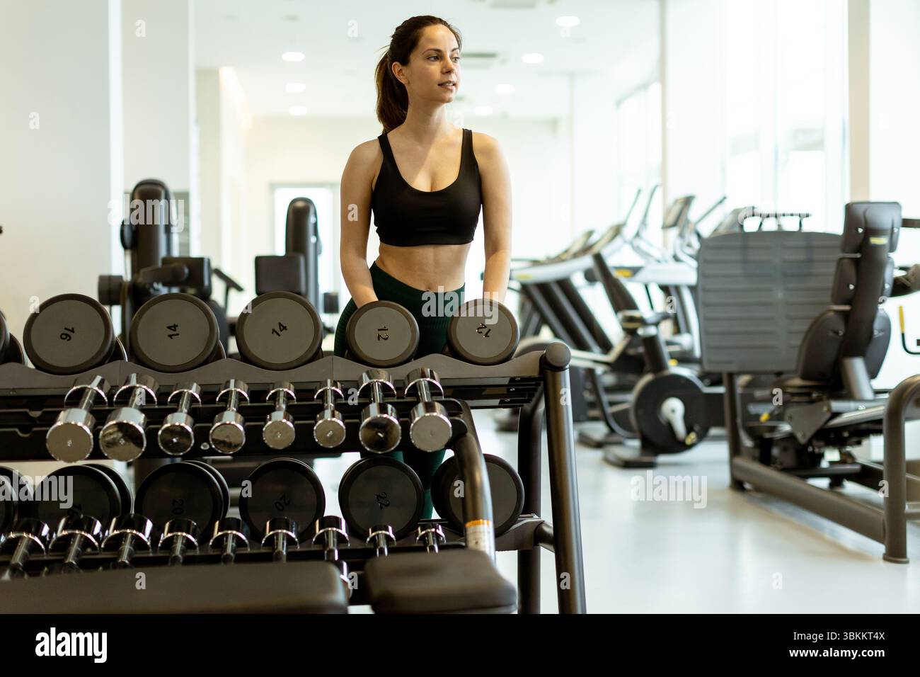 Una donna determinata si trova accanto a una cremagliera di manubri, pronta a migliorare la sua forza in un ambiente di palestra luminoso ed energico. Foto Stock