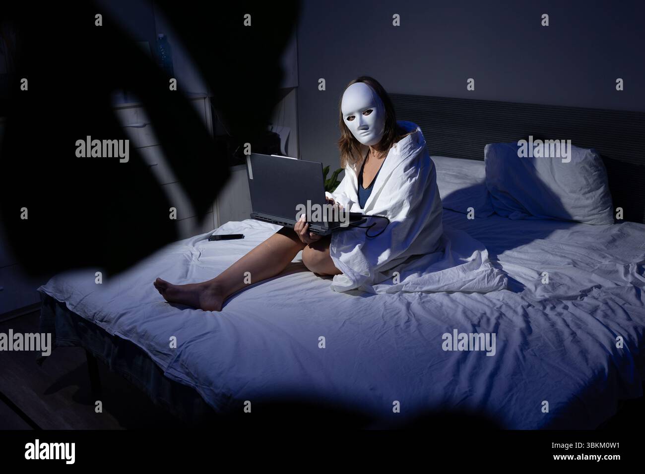 In una camera da letto poco illuminata, un individuo mascherato si siede su un letto con un notebook, sottolineando l'importanza della privacy digitale e la minaccia di frodi ai dati. Foto Stock