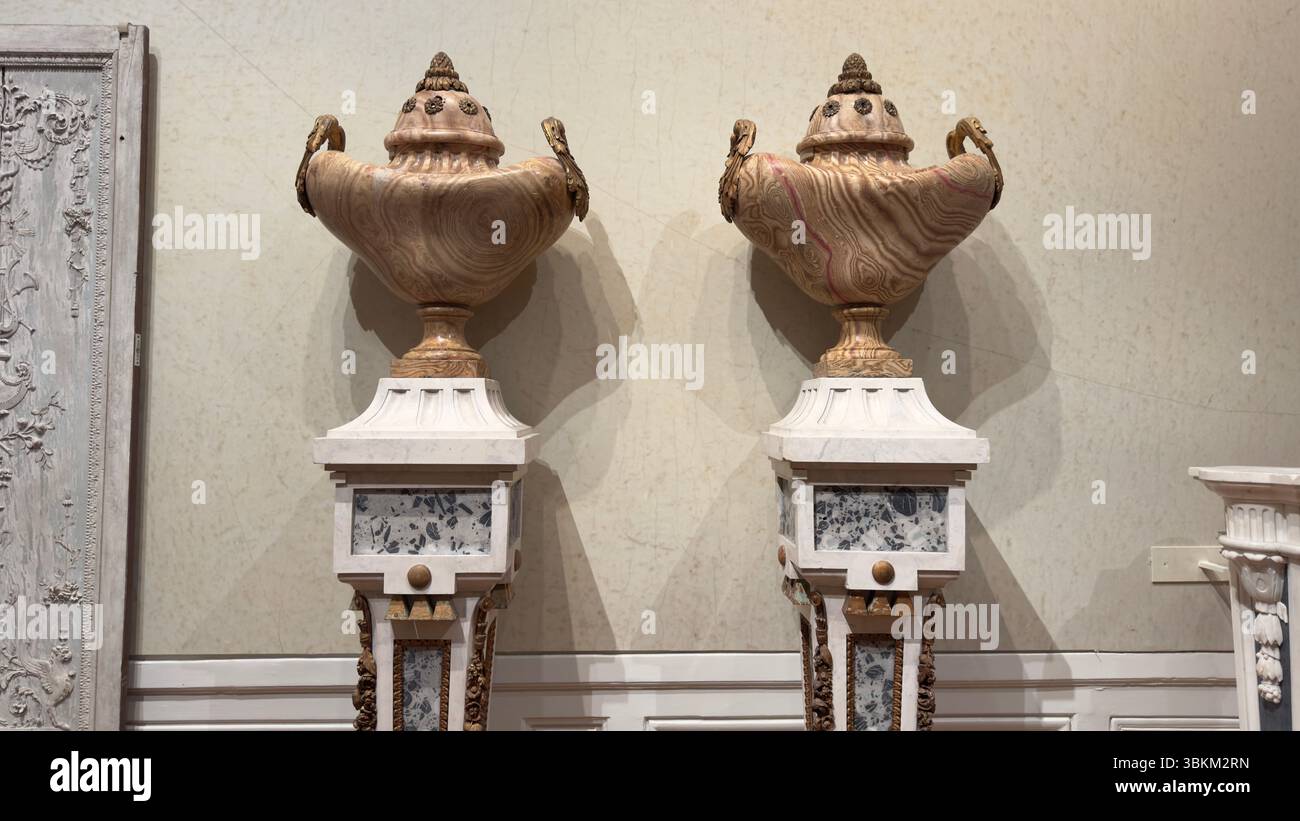 Due urne di pietra ornate si trovano in cima ai piedistalli di marmo contro una parete neutra. Le finiture e le incisioni dettagliate aggiungono interesse visivo. Foto Stock