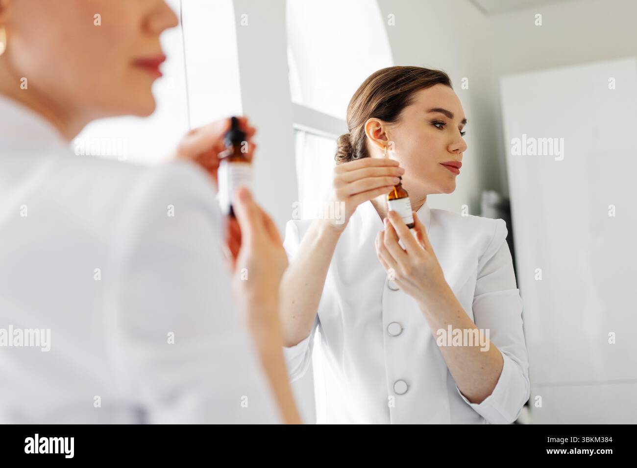 Donna in camice bianco che tiene in mano siero anti-invecchiamento e contagocce vicino al viso, davanti allo specchio in un centro di bellezza, che dimostra un prodotto per la cura della pelle professionale f Foto Stock
