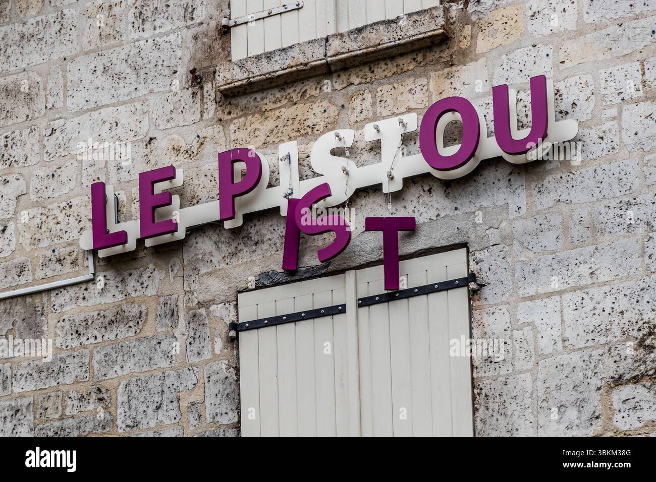 Un cartello danneggiato reca il nome di un negozio, Rue du Chat, Angoulême, Nouvelle-Aquitaine, Francia. Tre lettere del nome "Le Pistou" pendono solo dai cavi dell’illuminazione di fondo. Le lettere viola I, S e T sono scivolate dalla loro striscia di supporto bianca. Il cartello è appeso a una facciata in pietra chiara sopra una finestra con persiane in legno bianco. La città è una Città Creativa della Letteratura dell’UNESCO dal 2019. Foto Stock