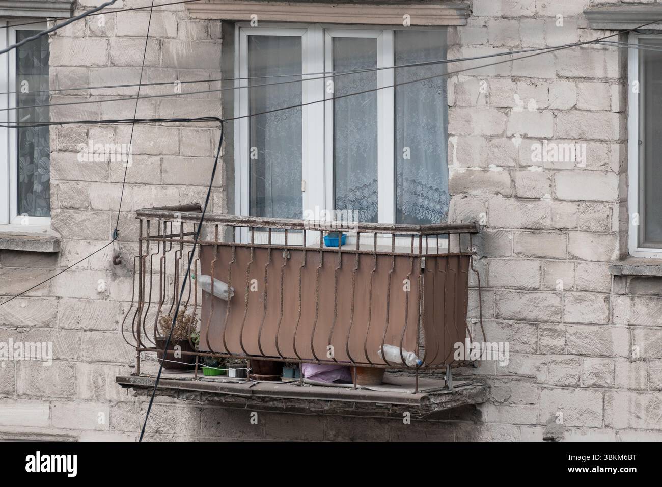 Un balcone urbano intempestivo presenta due piccioni arroccati su una ringhiera in metallo arrugginito, tra tende a rete e oggetti di uso quotidiano su un edificio in cemento Foto Stock