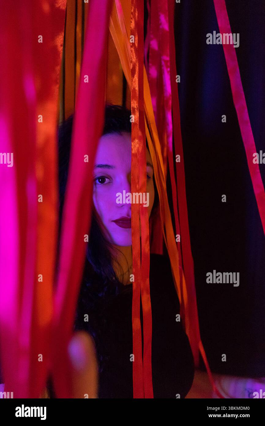 Una donna con un sottile sorriso sbircia attraverso una tenda di nastri vibranti, il suo volto illuminato da una luce viola tenue. La scena trasuda un senso di miste Foto Stock