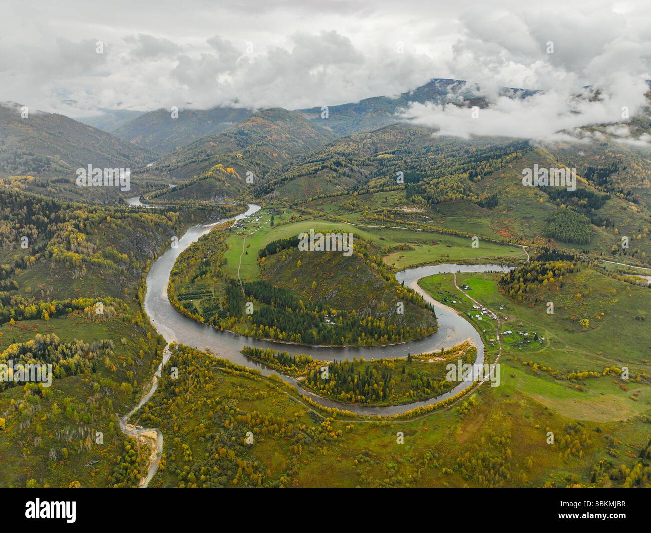 Una vista aerea di un fiume serpeggiante attraverso un paesaggio autunnale con edifici sparsi e montagne lontane sotto cieli nuvolosi Foto Stock