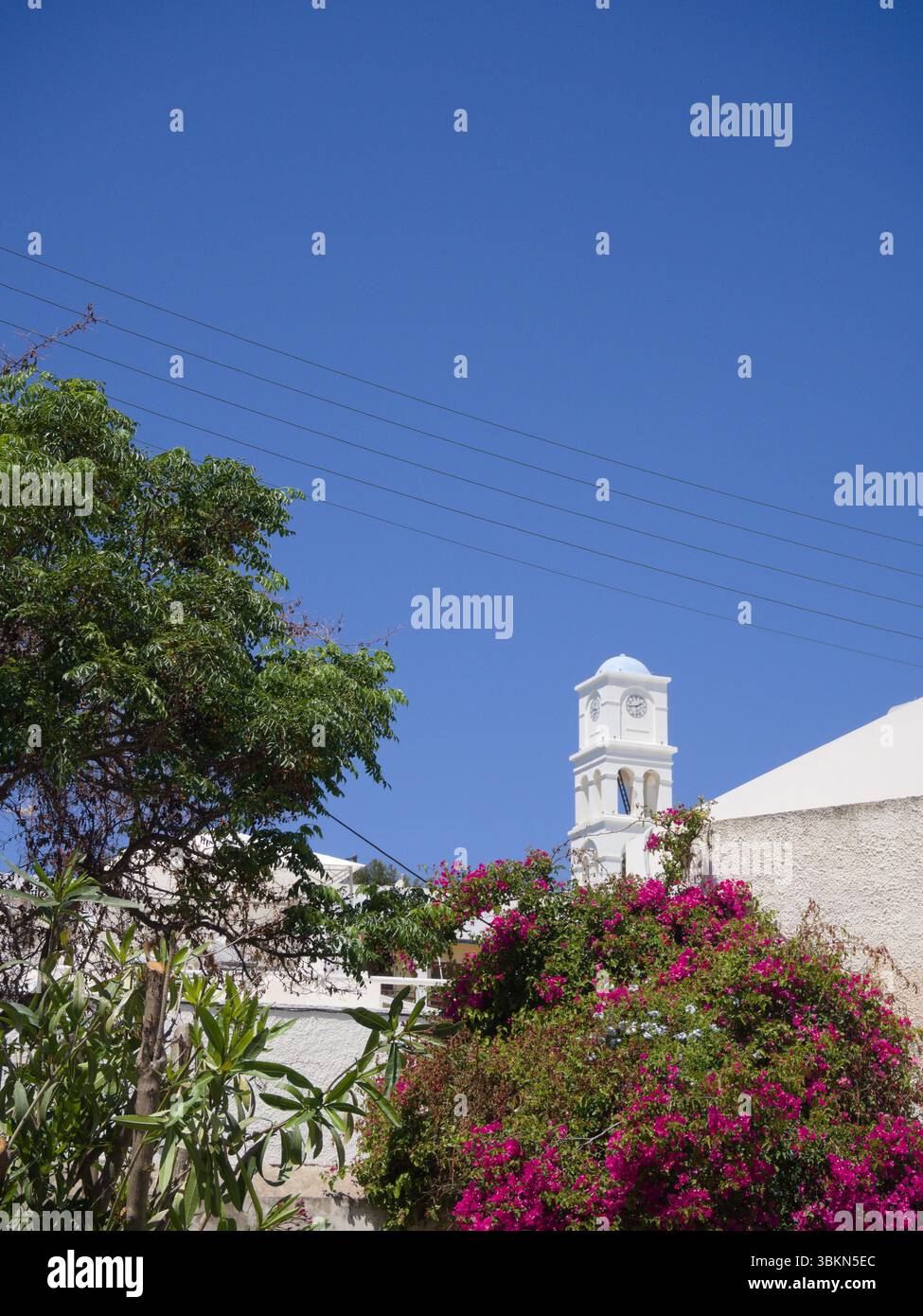 Torre bianca della chiesa con accenti blu chiaro in un villaggio mediterraneo circondato da bouganville, pini e vegetazione. Foto Stock