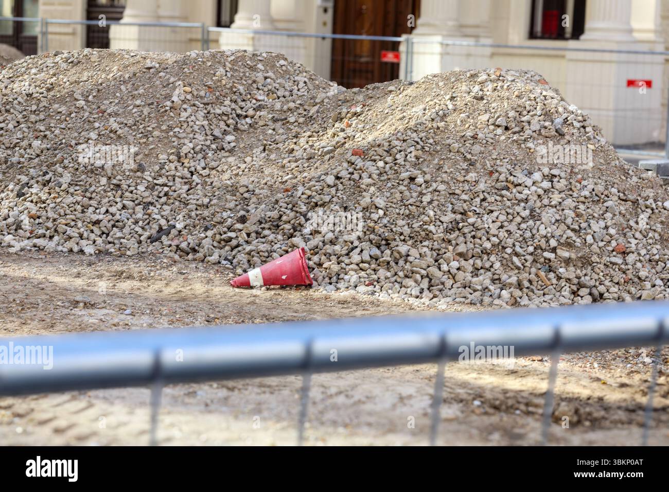 Scena di un cantiere con un grande mucchio di ghiaia o macerie sulla destra, un cono rosso stradale in primo piano e una strada con veicolo Foto Stock