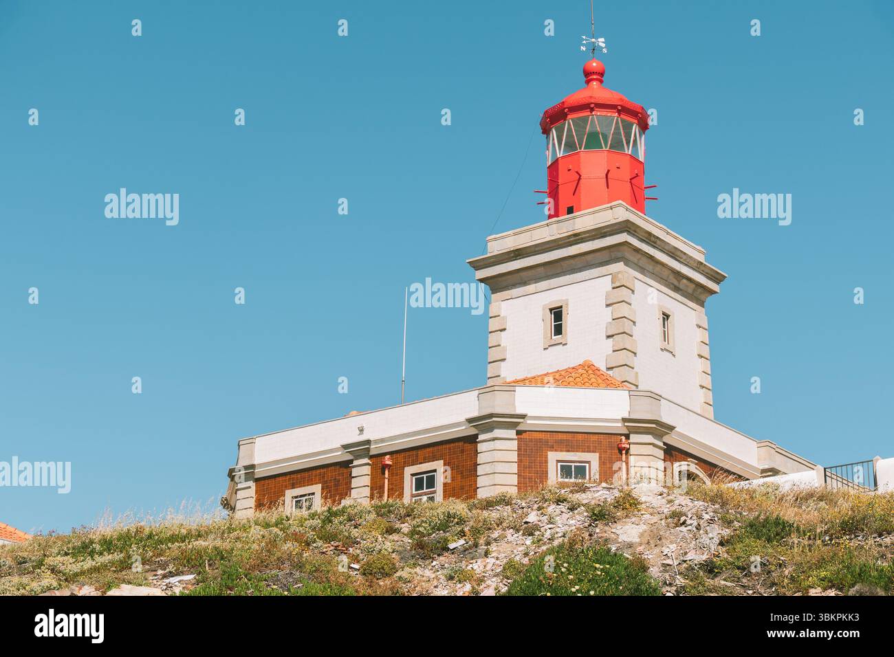 Il faro rosso brillante sorge su una collina rocciosa sotto il cielo azzurro nella regione di Lisbona, in Portogallo. Ideale per viaggi, turismo, navigazione e sicurezza marittima. Foto Stock