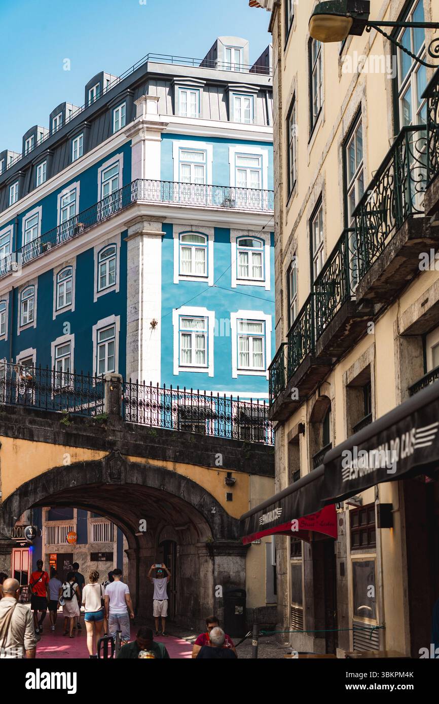 Vivace vista sulla strada di Lisbona caratterizzata da un suggestivo edificio blu, architettura storica e persone che camminano sotto un arco, perfetto per viaggi, turismo e temi di stile di vita urbano. Foto Stock