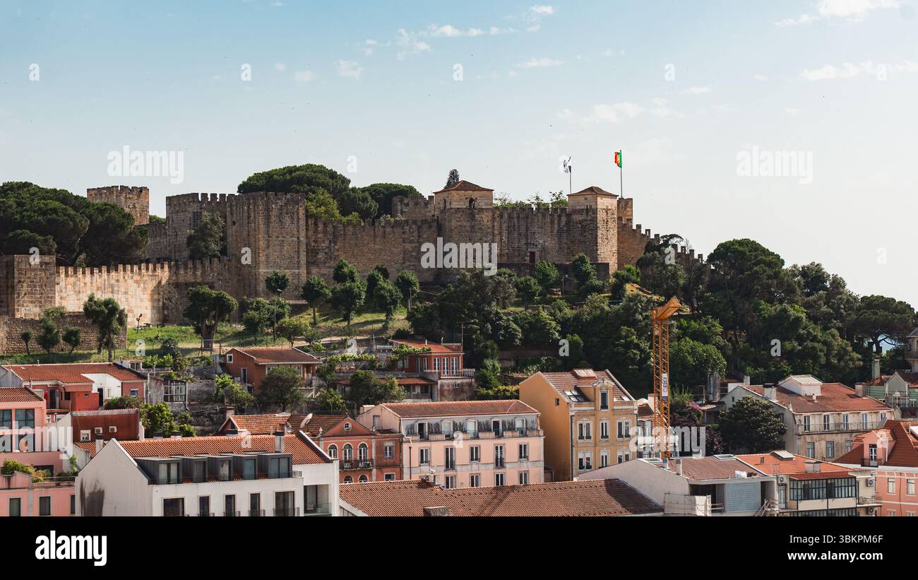 Vista panoramica del castello di Sao Jorge che si innalza sopra i vivaci tetti di Lisbona, circondato da vegetazione lussureggiante. Perfetto per viaggi, architettura e ispirazione turistica portoghese. Foto Stock