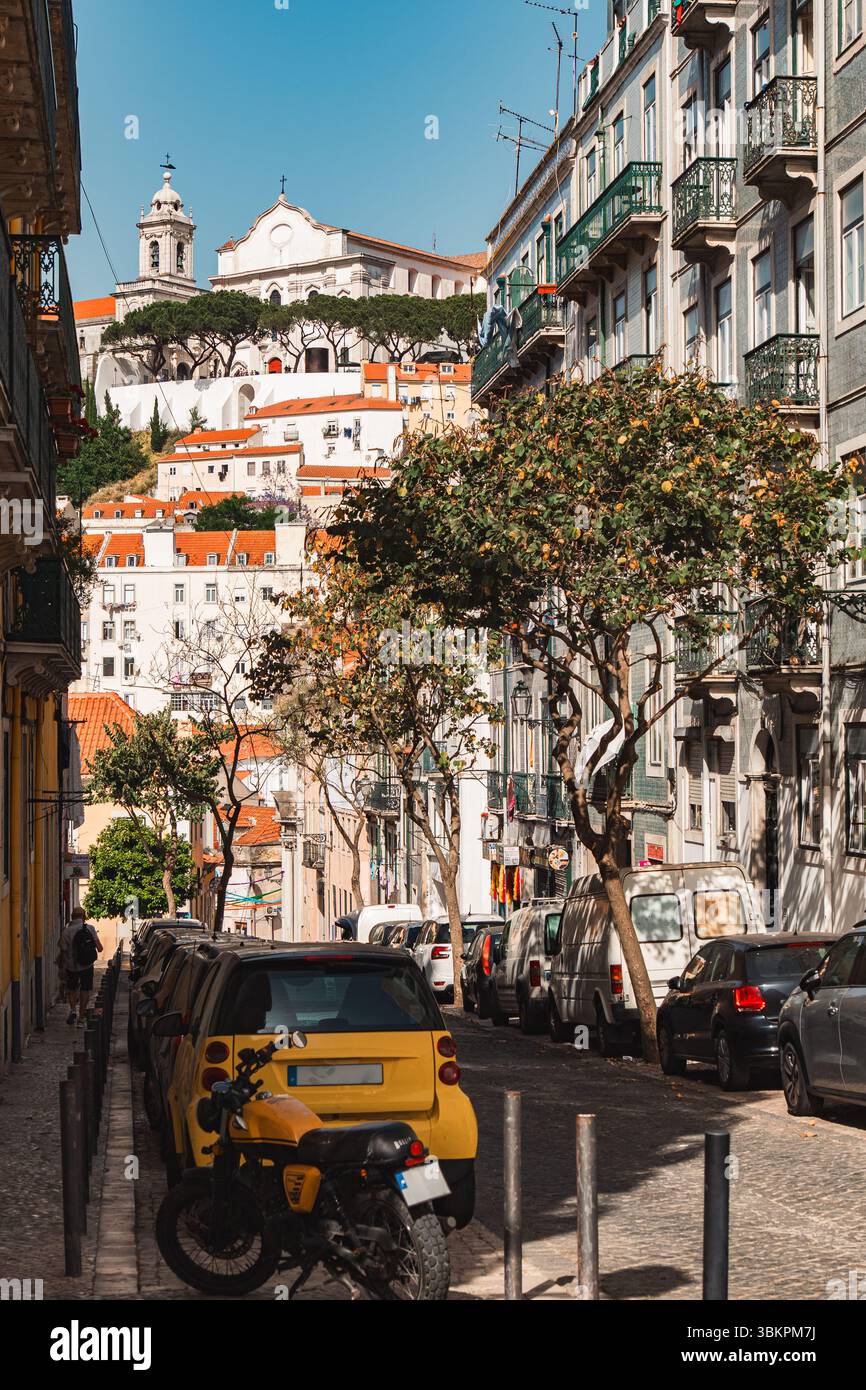Pittoresca strada di Lisbona caratterizzata da automobili vivaci, edifici tradizionali, alberi frondosi e una vista panoramica di una chiesa storica in cima a una collina sotto un cielo azzurro. Foto Stock