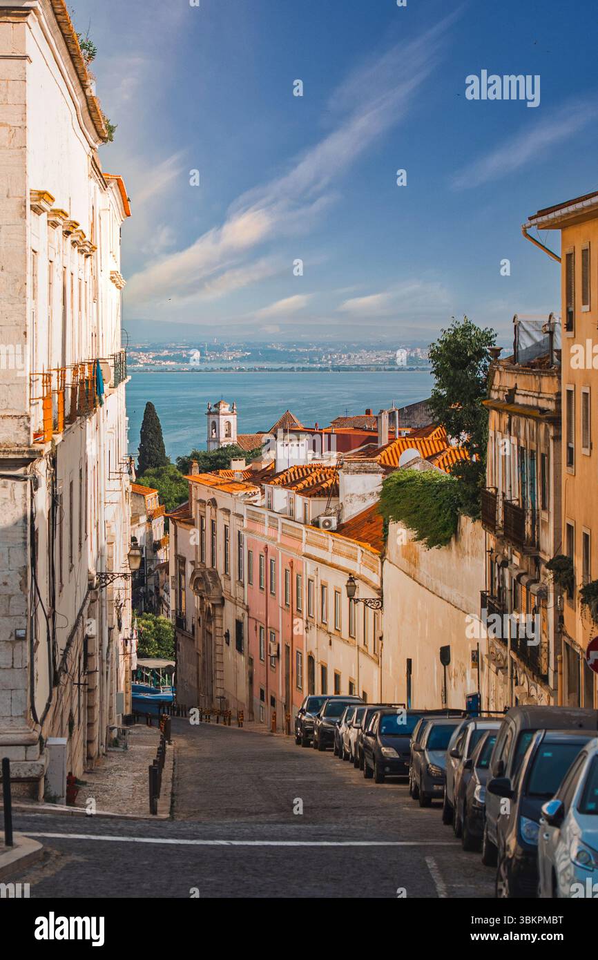 Affascinante strada di Lisbona caratterizzata da edifici colorati, auto parcheggiate e una splendida vista sul fiume Tago sotto un cielo blu brillante, perfetto per ispirarsi al viaggio. Foto Stock