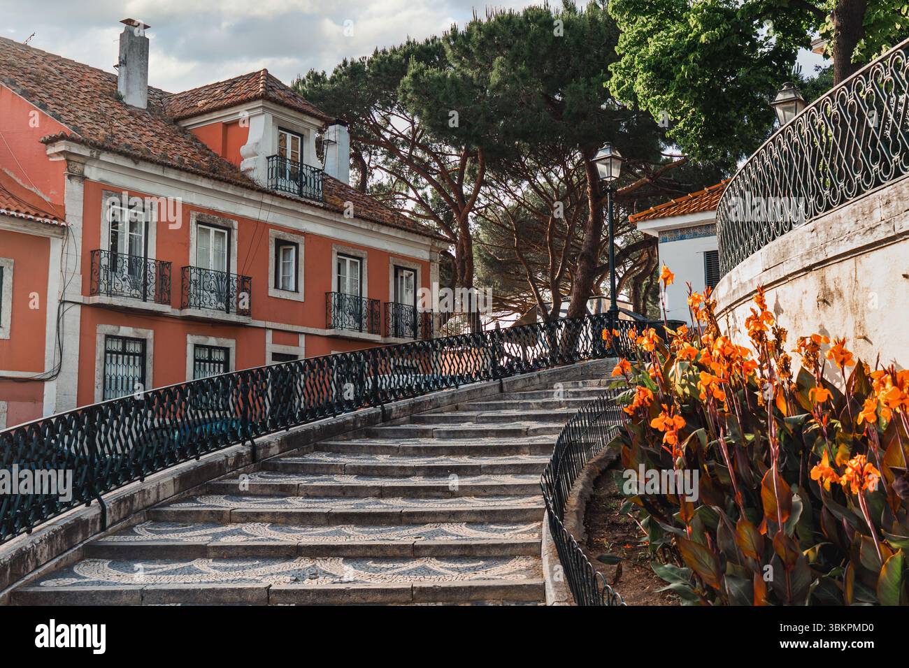 Pittoresca scalinata in pietra a Lisbona, Portogallo, caratterizzata da edifici tradizionali colorati, ringhiere ornate, vegetazione lussureggiante e vivaci fiori arancioni, perfetta per ispirare i viaggi e i temi del paesaggio urbano. Foto Stock