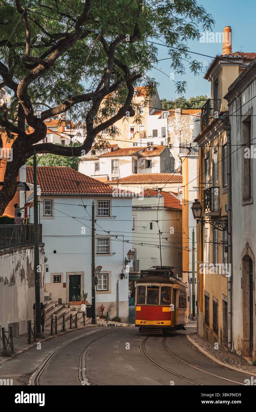 L'affascinante tram giallo viaggia attraverso le strette e tortuose strade del quartiere Alfama di Lisbona, circondato da case colorate, architettura tradizionale e lussureggianti rami di alberi. Foto Stock