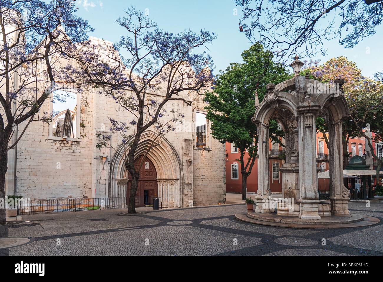 Splendida vista delle rovine del convento del Carmo e della fontana in pietra ornata in una tranquilla piazza di Lisbona, incorniciata da alberi di jacaranda e architettura storica, perfetta per ispirarsi ai viaggi. Foto Stock