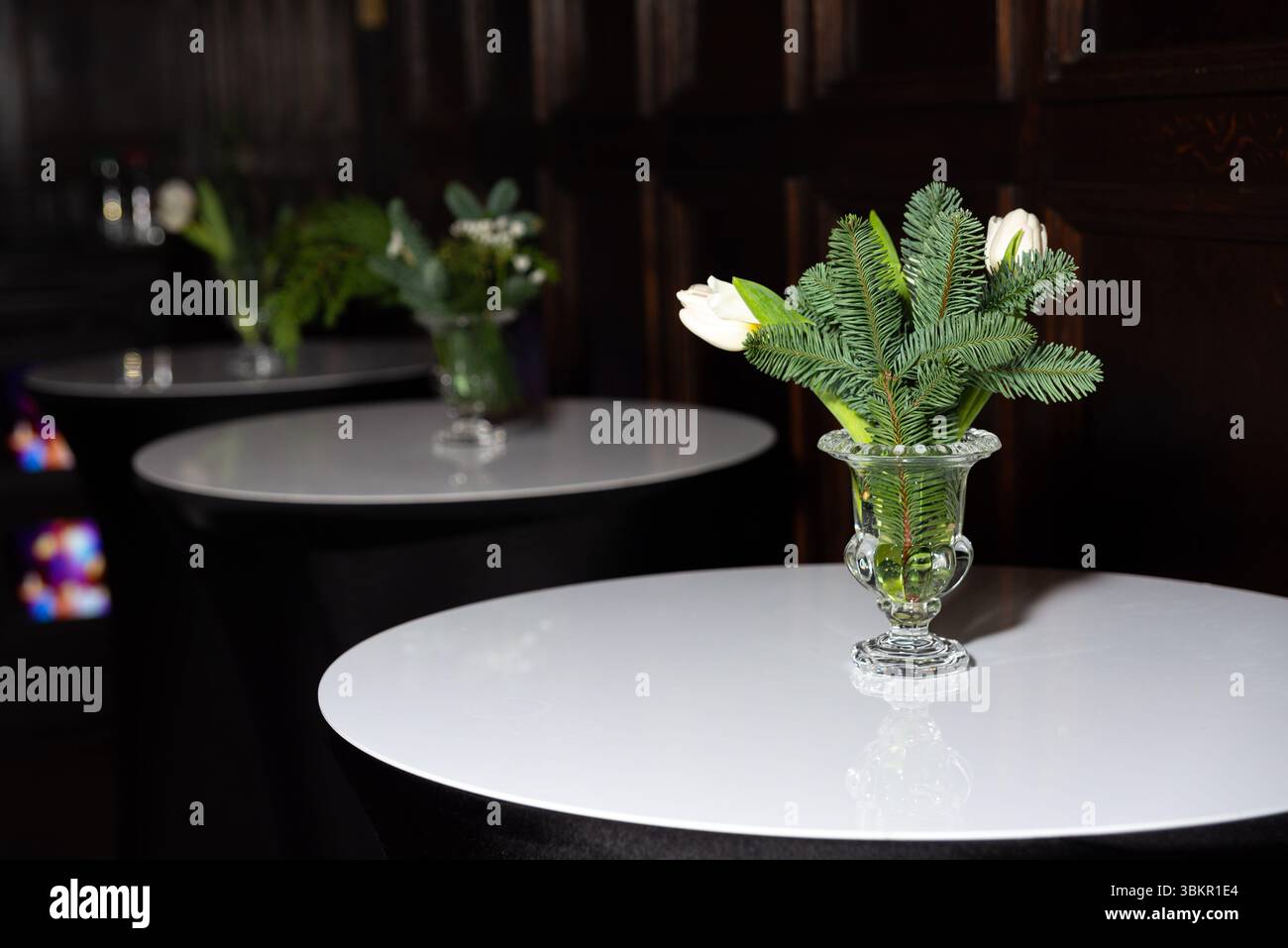 Vasi di vetro con tulipani bianchi e rametti di pino decorano moderni tavoli da cocktail bianchi in un ambiente scuro ed elegante. Foto Stock