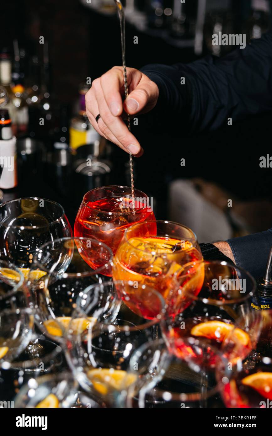 Primo piano di un barista che mescola cocktail Aperol spritz con fette d'arancia e ghiaccio in un ambiente da bar professionale. Foto Stock
